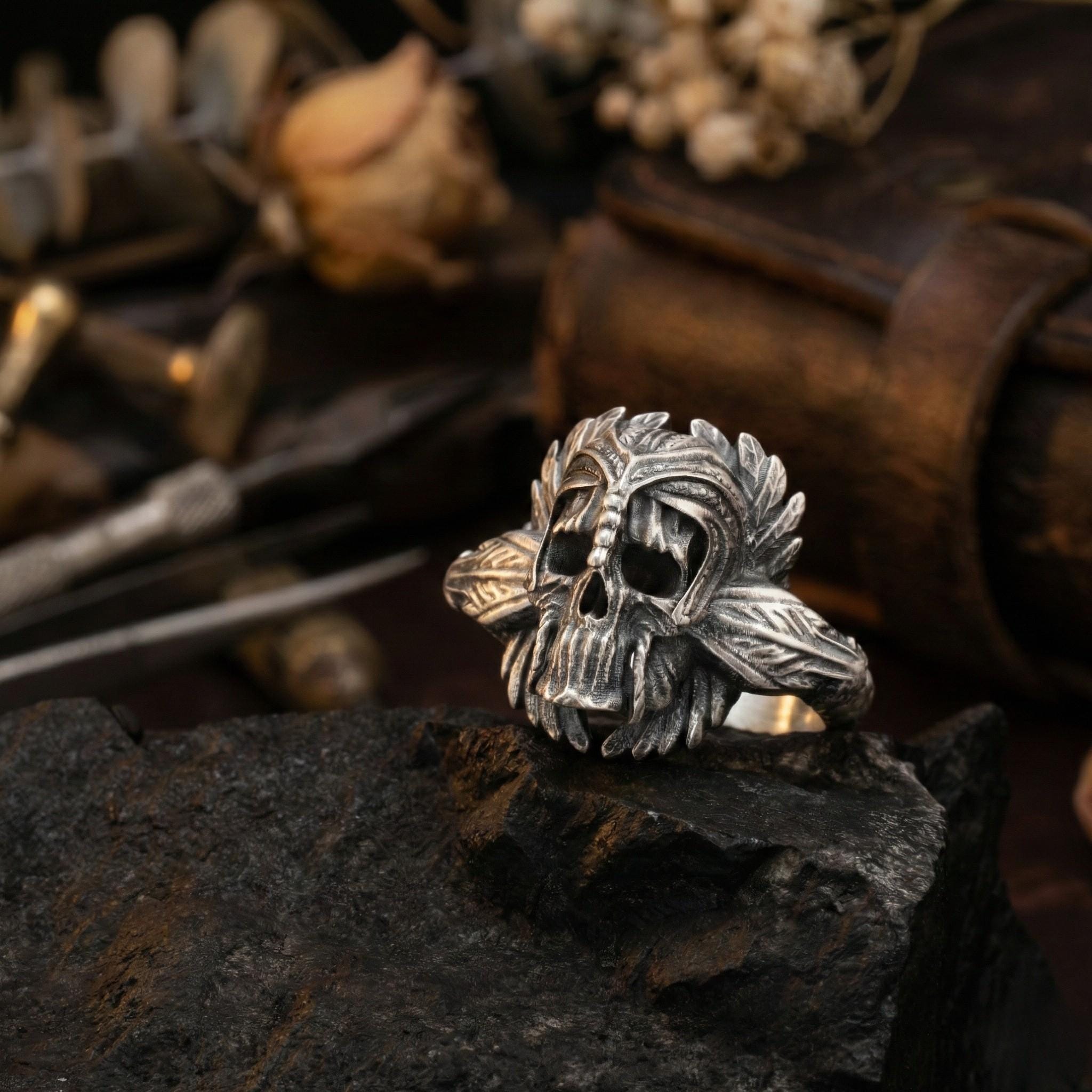 A silver skull ring with intricate leaf designs sits on a rock, surrounded by various objects such as a leather-bound book and a pair of scissors.